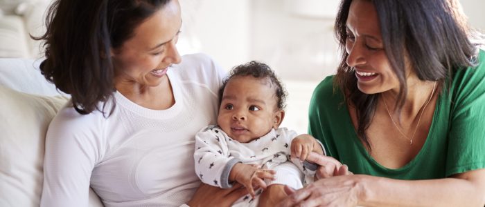 Millennial mixed race mother sitting in an armchair holding her three month old baby son, her mother kneeling beside them